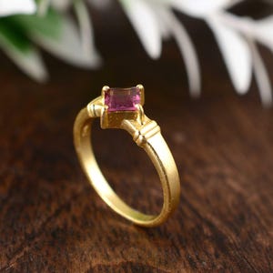 Op de afbeelding: Een goudkleurige ring met een rechthoekige, roze edelsteen. De ring heeft een eenvoudige band en een decoratieve zetting rond de steen. De edelsteen heeft een levendige roze kleur en de ring lijkt van goud te zijn.
