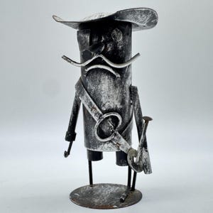 May include: A handcrafted metal sculpture of a pirate figure. The sculpture is painted in black and white, with a hat, mustache, and sword. The figure stands on a circular base, and the overall design has a rustic, vintage aesthetic.