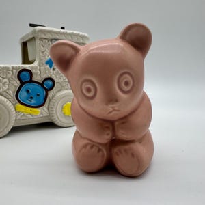 Op de afbeelding: Een vintage roze keramieken berenbeeldje met een lieve uitdrukking, zittend met gevouwen poten. Op de achtergrond een witte trein-vormige container met blauwe berendetails en bloemmotieven. Een charmant, nostalgisch decoratief stuk.