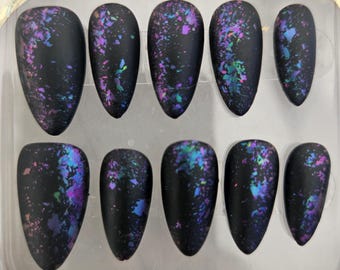 May include: A set of ten almond-shaped press-on nails with a matte black base finish. Each nail features a scattered, iridescent flake design in vibrant shades of purple, blue, and green, creating a galaxy-like effect across the dark surface.