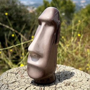 May include: A ceramic Moai statue in shades of gray and brown, with a prominent nose and facial features. The statue is displayed outdoors on a weathered wooden surface, with a blurred natural background.