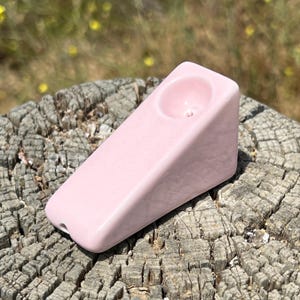 May include: A pink ceramic pipe with a smooth, glossy finish. The pipe has a triangular shape with a bowl at the top and a hole at the end. The pipe is resting on a weathered wooden surface.