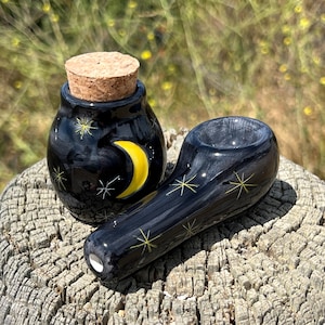 May include: A black ceramic pipe and a small jar with a cork stopper. The jar features a yellow crescent moon and star designs. The pipe is decorated with yellow star patterns.