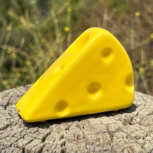 May include: A bright yellow, wedge-shaped cheese sculpture with several round holes. The cheese is sitting on a weathered, textured wooden surface, with a blurred green background.