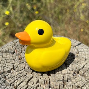 May include: A bright yellow rubber duck with an orange beak and a black eye. The duck is sitting on a weathered wooden surface, with a blurred green background. The duck is a classic toy.