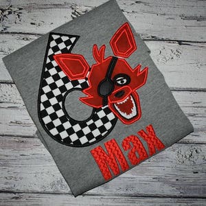 May include: A gray t-shirt with a black and white checkered number six and a red fox character with a black eye patch. The name Max is embroidered in red below the number.