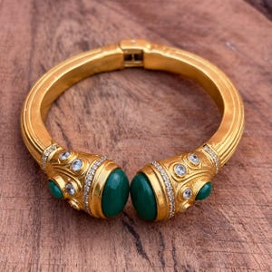 Könnte beinhalten: Ein goldfarbenes Klapparmband mit zwei großen, glatten, dunkelgrünen Cabochon-Steinen an den Enden. Die Endstücke sind mit kleineren, runden, hellblauen Steinen und Reihen winziger, klarer Kristalle auf der Metalloberfläche verziert.