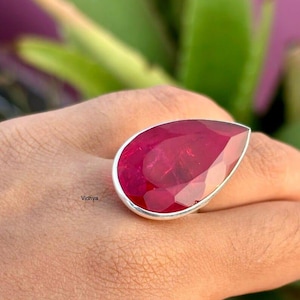 Puede incluir: Un anillo con una gran piedra preciosa roja rubí en forma de pera, engastada en una banda plateada. La piedra está facetada, captando la luz y mostrando un color profundo y rico. El anillo es una pieza llamativa.