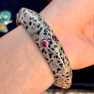 Op de afbeelding: Een gespikkelde armband met zwarte vlekken op een lichte achtergrond, versierd met drie rode edelstenen. De armband wordt om een pols gedragen, wat het ontwerp en het kleurcontrast benadrukt. De edelstenen zijn in zilver gezet.