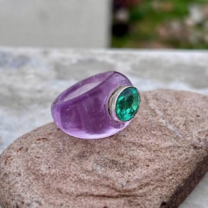 May include: A translucent light purple ring featuring a smooth, rounded band. The ring is set with a single, oval-shaped, faceted green gemstone held in a silver-toned metal bezel, creating a distinct contrast between the purple and green colors.