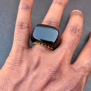 以下が含まれることがあります： 滑らかで光沢のあるダークブラウンの長方形のトップを備えたリング。リングのベースは質感のあるゴールドトーンの金属素材で構成されており、バンドの暗くしっかりとした上部と対照的です。