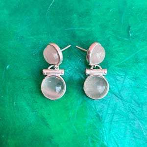 Pode incluir: Um par de brincos de prata com um design de dois n&iacute;veis. Cada brinco apresenta uma pequena pedra rosa claro em forma de cora&ccedil;&atilde;o no topo e uma pedra transl&uacute;cida redonda maior na parte inferior. Em um fundo verde.