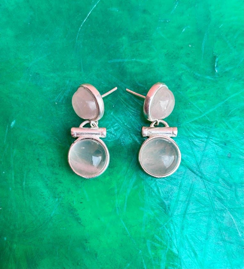 Pode incluir: Um par de brincos de prata com um design de dois n&iacute;veis. Cada brinco apresenta uma pequena pedra rosa claro em forma de cora&ccedil;&atilde;o no topo e uma pedra transl&uacute;cida redonda maior na parte inferior. Em um fundo verde.