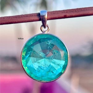 以下が含まれることがあります： 銀色の金属フレームにセットされた、ファセット加工された表面を持つ丸いターコイズ色の宝石ペンダント。ペンダントは銀色のベイルから吊り下げられています。背景はぼやけており、ピンクと緑の色合いがあります。