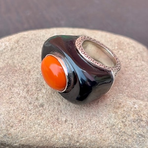May include: A chunky ring featuring a large, oval orange gemstone set in a black, glossy base. The band is silver with a decorative pattern around the edge. The ring is displayed on a textured, light brown surface.