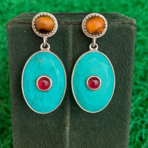 以下が含まれることがあります： 銀色のイヤリングのペア。各イヤリングは、上部に丸い琥珀色の石があり、中央に赤い石が付いたターコイズ色の楕円形に接続されています。イヤリングは緑色の背景に表示されています。