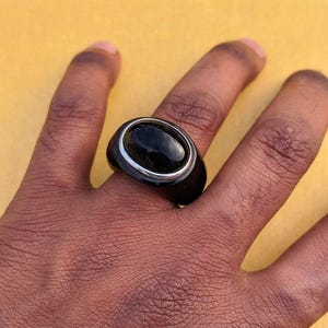 以下が含まれることがあります： 大きな楕円形の黒い石が銀色のベゼルにセットされた黒い指輪。指輪は太い黒いバンドが付いています。指輪は人の指に表示されています。