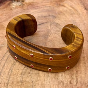 May include: A wide, open-cuff bracelet made of tiger's eye stone. The bracelet is a warm brown color with darker brown stripes. Small, round red stones are set into the bracelet with gold-colored metal settings.