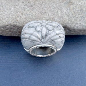 以下が含まれることがあります： 白とグレーの彫刻が施された石の指輪、花の模様。 指輪は、ビーズの縁が付いた銀色のバンドが付いています。 石はマーブル模様で、青い背景にセットされています。