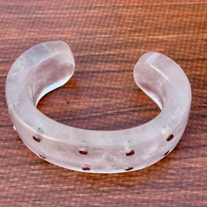 Könnte beinhalten: Ein klarer, gebogener Armreif mit kleinen, runden, roten Akzenten. Das Armband besteht aus einem durchscheinenden Material und hat eine glatte, polierte Oberfläche. Das Armband wird auf einer Holzoberfläche gezeigt.