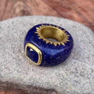 May include: A thick, deep blue lapis lazuli ring featuring a gold-toned metal inlay around the inner band with a sunburst-style edge. A single, square-cut blue gemstone is set into the front of the band, framed by a smooth, gold-toned metal border.