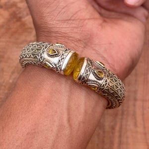 Könnte beinhalten: Silberfarbenes Armband mit aufwendigen geometrischen Mustern und gelben Edelsteinakzenten. Das Armband hat ein Scharnierdesign mit einem zentralen gelben Edelsteinelement. Das Armband wird am Handgelenk getragen und zeigt sein Design und seine Handwerkskunst.