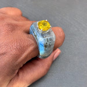 以下が含まれることがあります： 磨き上げられた仕上げと青い虹色の輝きが見えるグレーのラブラドライト石で作られたチャンキーなリング。リングの上部には、4本の爪を持つ銀色の金属製の台座にしっかりと固定された、目立つ正方形カットの鮮やかな黄色の宝石があしらわれています。