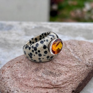 May include: A ring featuring a thick, cream-colored band with a black spotted pattern resembling dalmatian fur. The band is set with a single, round, faceted orange gemstone held in a silver-toned metal setting that protrudes from the side.