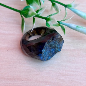 Op de afbeelding: Een gefacetteerde, donkerblauwe en bruine edelsteenring met een ruwe, getextureerde rand. De ring ligt op een lichtroze oppervlak, met groene en witte decoratieve takjes op de achtergrond. De facetten van de ring vangen het licht.
