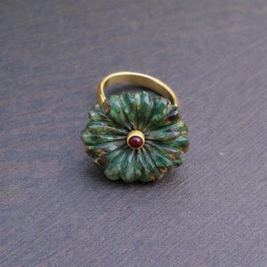 Könnte beinhalten: Ein goldener Ring mit einem geschnitzten grünen Stein in Blütenform und einem kleinen roten Edelstein in der Mitte. Der Ring hat ein goldenes Band und die Blume detaillierte Petalenschnitzereien.
