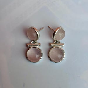 Pode incluir: Um par de brincos de prata. Cada brinco apresenta uma pequena pedra redonda rosa claro no topo, conectada a uma pedra redonda rosa claro maior na parte inferior. Design simples e elegante.