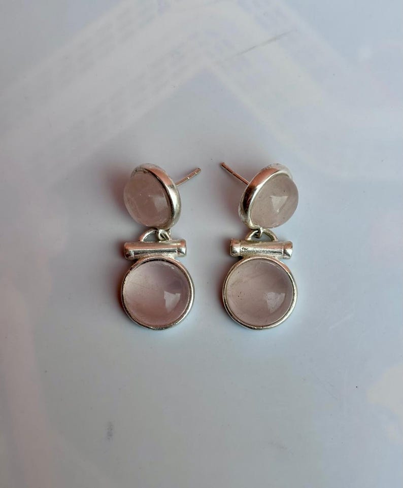 Pode incluir: Um par de brincos de prata. Cada brinco apresenta uma pequena pedra redonda rosa claro no topo, conectada a uma pedra redonda rosa claro maior na parte inferior. Design simples e elegante.