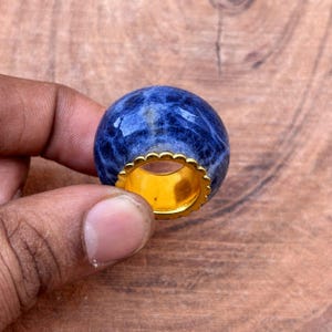 以下が含まれることがあります： 金色のスカラップバンドが付いた、青いソーダライトの宝石リング。 リングの宝石は、白の大理石模様の深い青色です。 リングは木製の背景に対して手で持たれています。