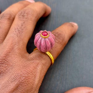 以下が含まれることがあります： 小さなカボチャや花のような形をした、彫刻が施されたリブ状のピンク色の宝石があしらわれたゴールドリング。ピンク色の石の中央には、ゴールドのベゼルにセットされた滑らかで丸い深紅の石が配置されています。