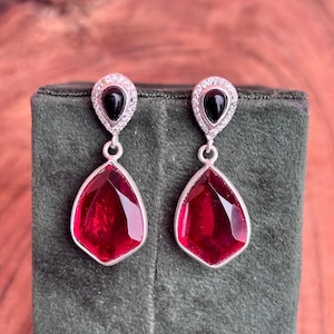 May include: A pair of silver-toned drop earrings. Each earring features a teardrop-shaped, faceted, deep red gemstone, suspended below a smaller black stone surrounded by clear crystals. The earrings are displayed on a dark green surface.