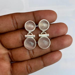 Pode incluir: Um par de brincos de prata com um design de c&iacute;rculo duplo. Cada brinco tem duas pedras redondas rosa claro emolduradas em uma moldura de metal prateado. Os brincos s&atilde;o conectados por uma pequena barra de prata.