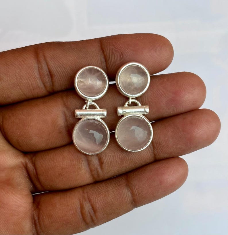 Pode incluir: Um par de brincos de prata com um design de c&iacute;rculo duplo. Cada brinco tem duas pedras redondas rosa claro emolduradas em uma moldura de metal prateado. Os brincos s&atilde;o conectados por uma pequena barra de prata.