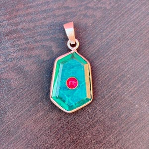 Puede incluir: Un colgante con un diseño geométrico. El colgante presenta una gran piedra preciosa de color verde esmeralda con una pequeña piedra roja redonda en el centro. El colgante está engastado en un metal dorado y tiene un lazo para una cadena.