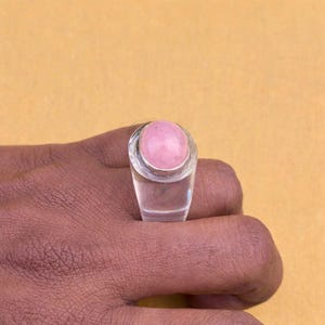 以下が含まれることがあります： 大きな楕円形のピンク色の宝石が付いた、透明な長方形のリング。 リングは黄色の背景に対して手にセットされています。 宝石は薄いピンク色です。