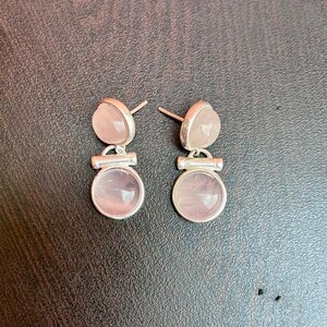 Pode incluir: Um par de brincos de press&atilde;o com pedras de quartzo rosa. Cada brinco tem uma pedra superior pequena e arredondada e uma pedra inferior circular maior, ambas em um tom rosa suave. As pedras s&atilde;o fixadas em metal prateado.