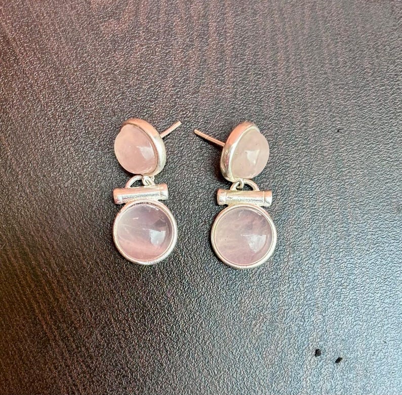 Pode incluir: Um par de brincos de press&atilde;o com pedras de quartzo rosa. Cada brinco tem uma pedra superior pequena e arredondada e uma pedra inferior circular maior, ambas em um tom rosa suave. As pedras s&atilde;o fixadas em metal prateado.