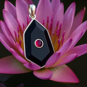 May include: A silver pendant with a black, geometric gemstone and a small, red center stone. The pendant hangs against a backdrop of a pink water lily, with dark green and black water in the background.