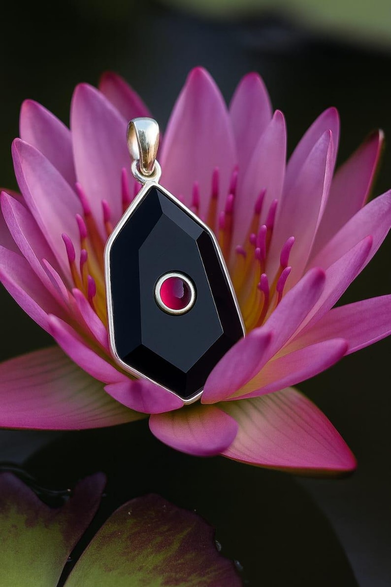 May include: A silver pendant with a black, geometric gemstone and a small, red center stone. The pendant hangs against a backdrop of a pink water lily, with dark green and black water in the background.