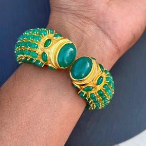 May include: Gold-toned cuff bracelet with large oval green gemstones and smaller green stones. The bracelet has a hinged design and is adorned with multiple emerald-green stones. The bracelet is on a person's wrist.