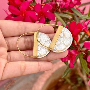 Könnte beinhalten: Goldfarbene Creolen mit einer weißen, floral gemusterten Scheibe und einem goldenen Balken. Die Ohrringe werden in einer Hand gehalten, mit leuchtend rosa Blumen im Hintergrund. Ein stilvolles Accessoire.