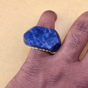 以下が含まれることがあります： 深い青色で白いマーブル模様が入った大きなファセットカットの石があしらわれた指輪。石はゴールドトーンの金属製バンドにセットされており、石座の縁にはテクスチャー加工されたビーズ状または刻み目のあるディテールが施されています。