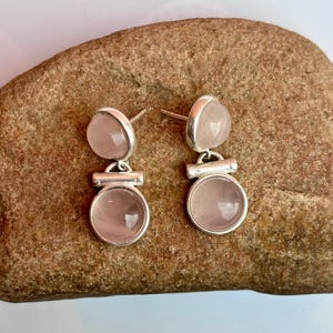 Pode incluir: Um par de brincos de prata com pedras de quartzo rosa. Cada brinco tem uma pedra inferior redonda e uma pedra superior menor e arredondada. Os brincos s&atilde;o exibidos em uma superf&iacute;cie marrom texturizada.