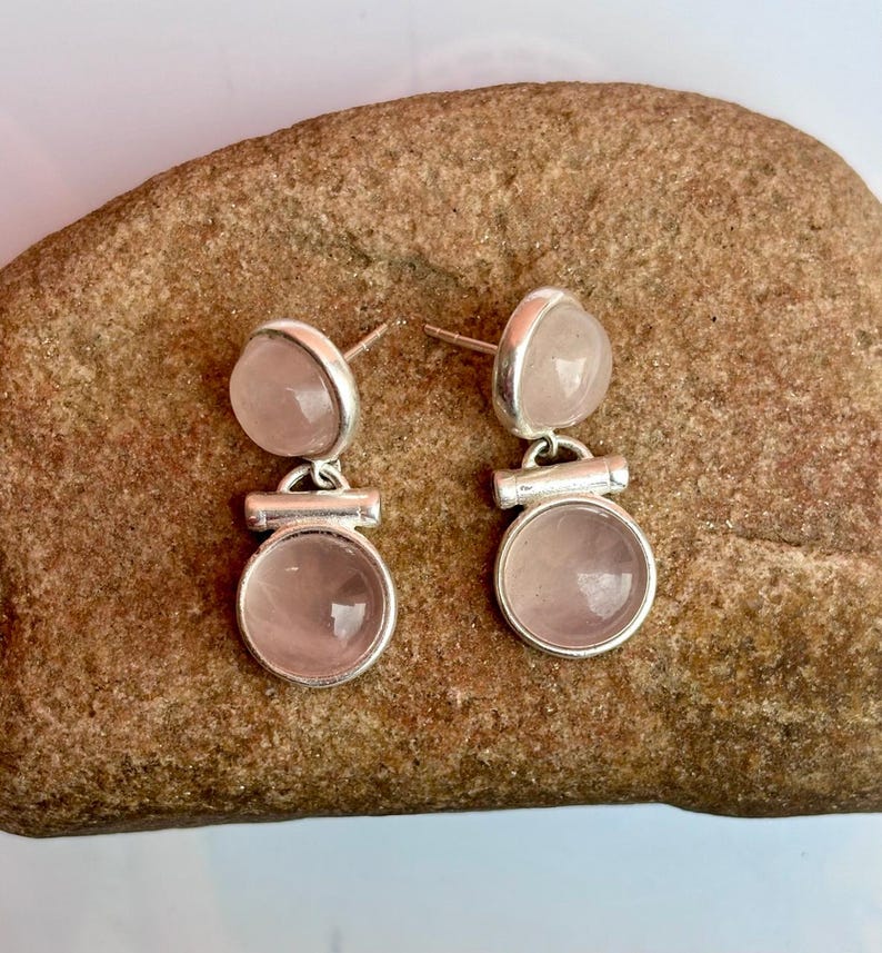 Pode incluir: Um par de brincos de prata com pedras de quartzo rosa. Cada brinco tem uma pedra inferior redonda e uma pedra superior menor e arredondada. Os brincos s&atilde;o exibidos em uma superf&iacute;cie marrom texturizada.