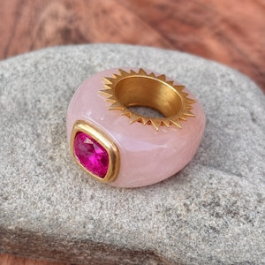 Pode incluir: Um anel robusto com uma banda de resina lisa em rosa pálido e uma incrustação de metal dourado em forma de raios de sol na borda interna. Uma pedra preciosa magenta quadrada e facetada está engastada numa moldura de metal dourado.