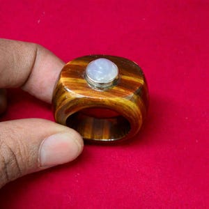以下が含まれることがあります： 虎の目のバンドと、銀色のベゼルにセットされた淡い青色の丸い宝石を特徴とする手作りのリング。 リングのバンドは、茶色と金色の温かみのあるストライプ模様を示し、ユニークで人目を引くアクセサリーです。